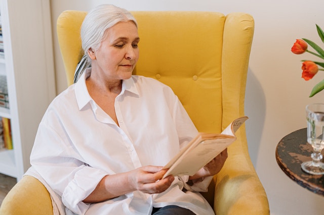 An older woman sitting on a bright yellow armchair is reading a book.