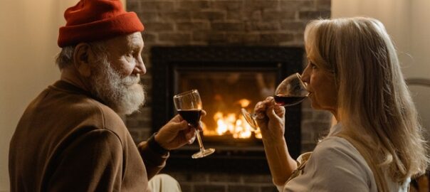 An elderly couple sitting on a dark brown leather couch and sipping wine in front of a fireplace.