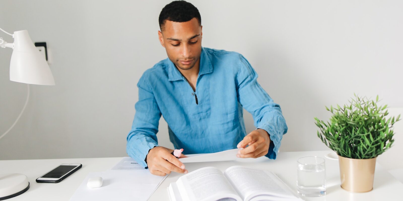 A man sitting at a white desk looking at a piece of paper with an open book in front of him.